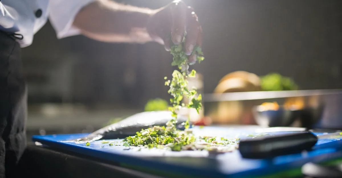 Main de chef préparant des légumes verts hachés sur une planche à découper dans une cuisine.