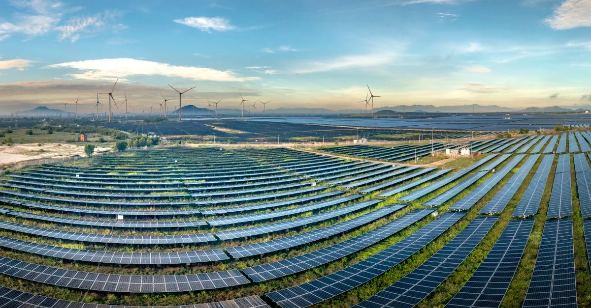 Vaste centrale solaire avec des éoliennes en arrière-plan, sous un ciel bleu azur.
