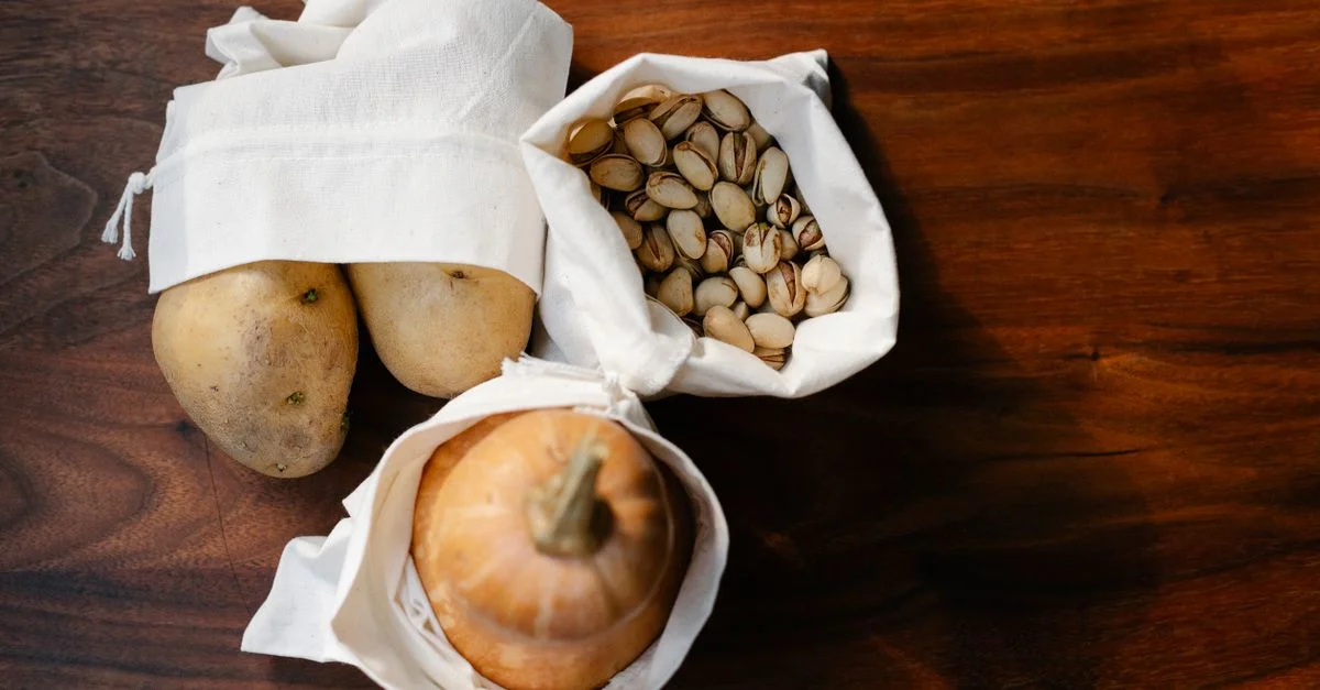 Vue du dessus de sacs réutilisables éco-responsables remplis de pommes de terre, de pistaches et d'une citrouille sur une table en bois.